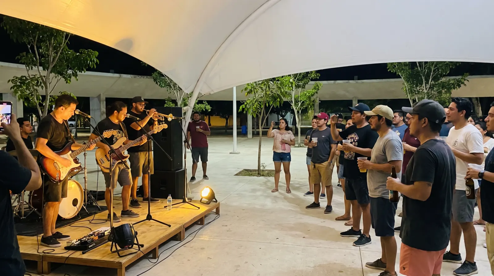 Banda tocando en un escenario bajo una carpa de noche, con el público de pie grabando con el celular